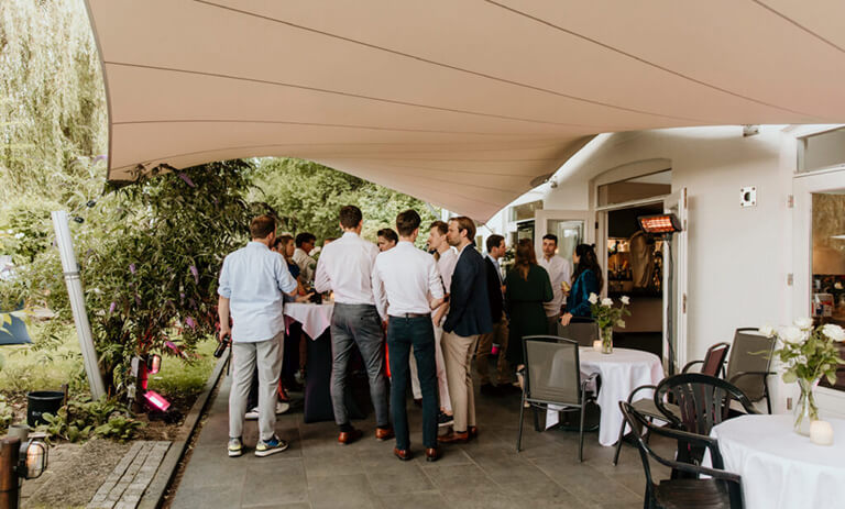 Group of people mingling at an outdoor reception under a tan canopy, around standing tables with chairs nearby and greenery in view.