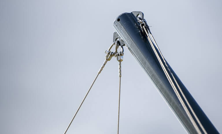Close-up of a sailboat masthead with rigging lines and hardware against a pale blue sky, showing cables and fittings at the top of the mast.