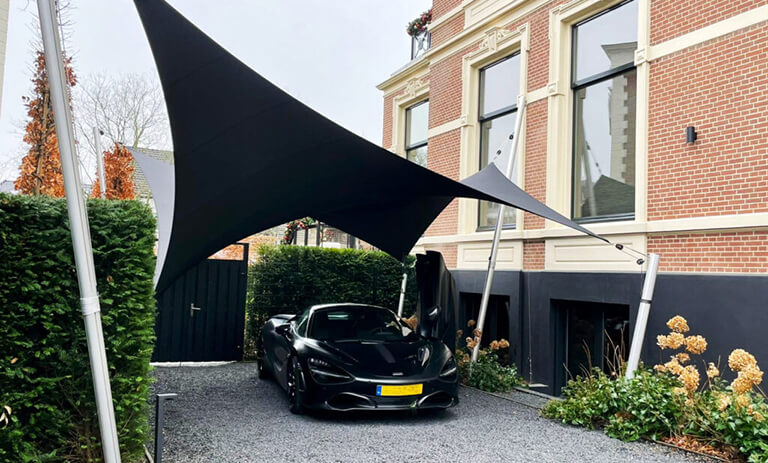Black sports car parked in a gravel driveway under a large black fabric shade sail beside a brick building.